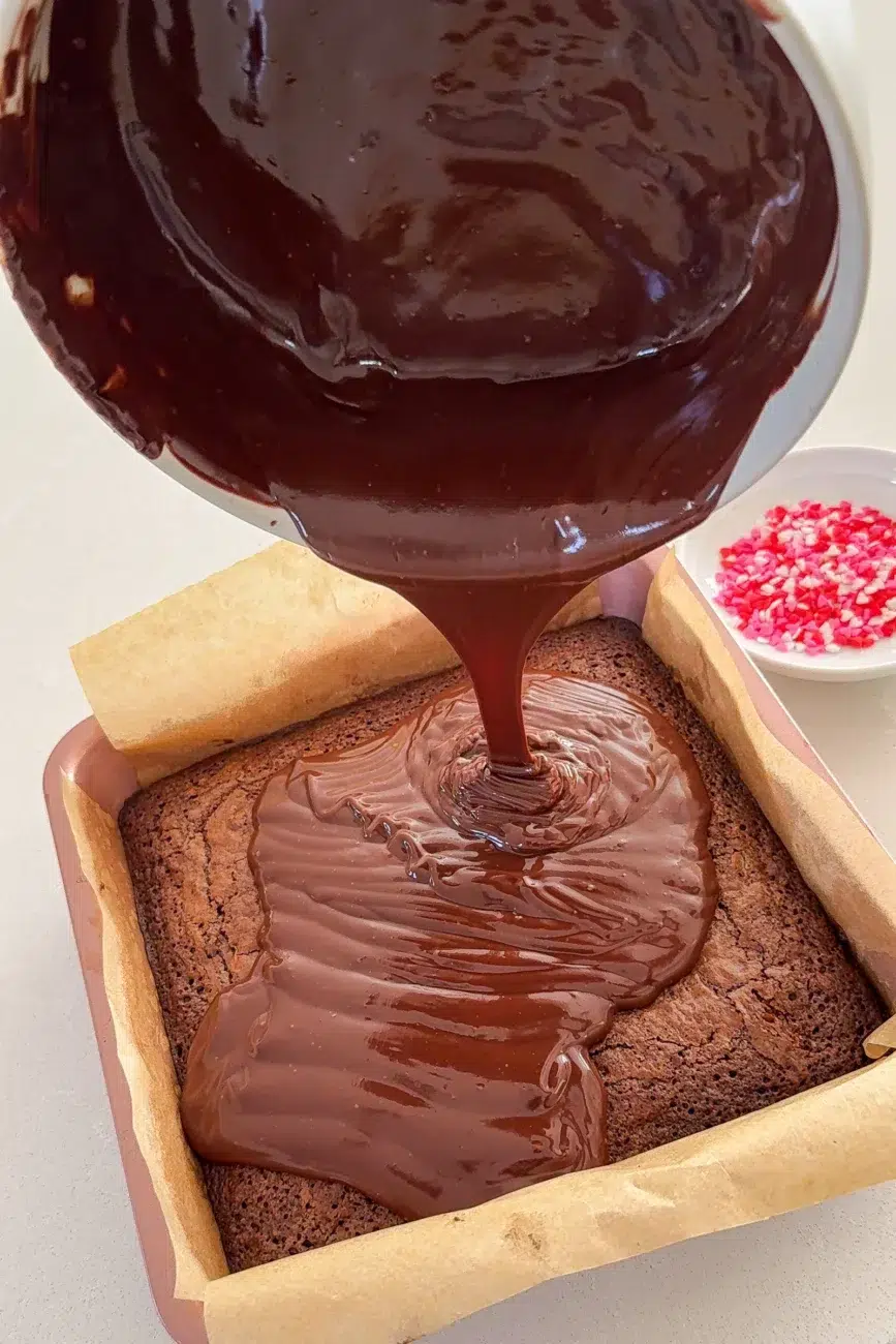 Chocolate ganache being poured onto cooled chocolate brownie in tin