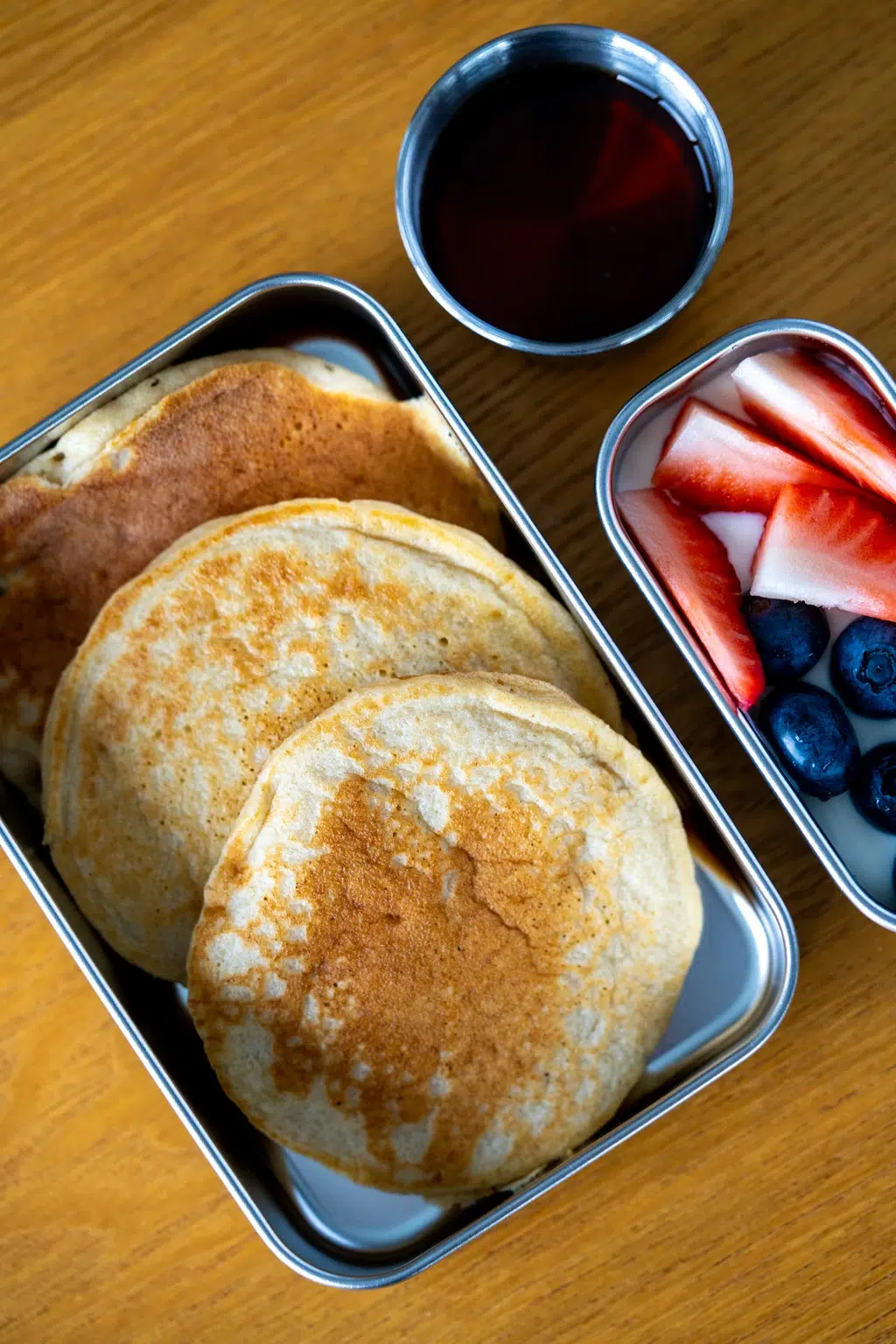 Bird’s eye view pancakes in stainless steel lunchbox with fruit and maple syrup on side