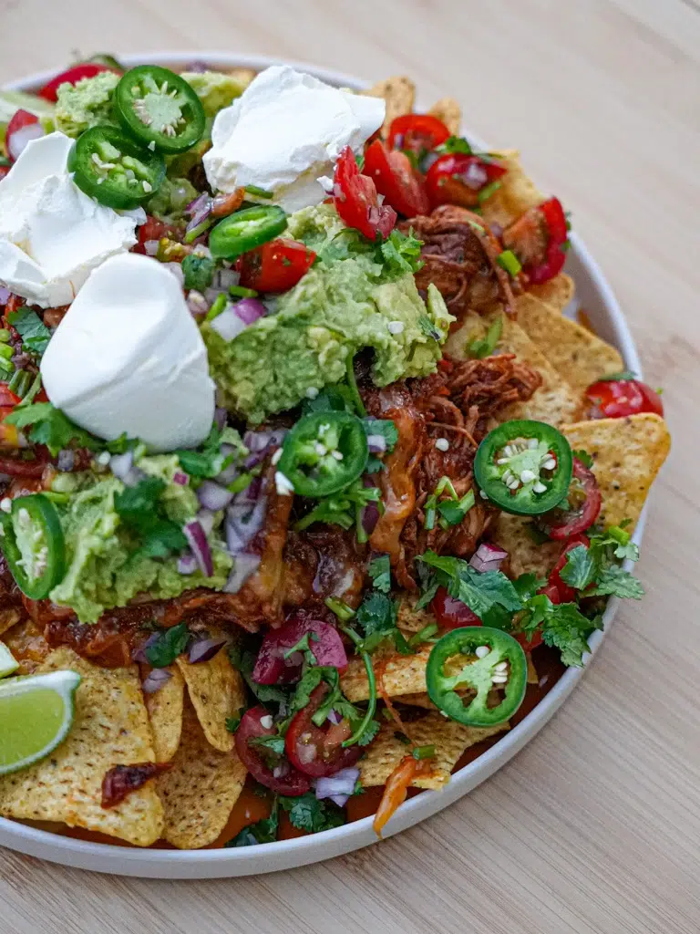 A bowl of crispy nachos loaded with pulled chicken, avocado, jalapeños, sour cream, tomatoes, and fresh cilantro