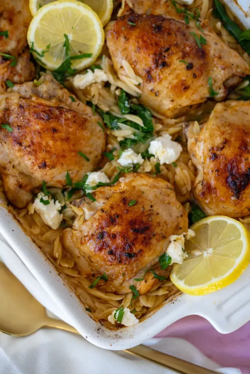 One pan Greek chicken with golden-brown crispy thighs, crumbled feta, lemon slices, and orzo pasta in a white baking dish, garnished with fresh herbs.