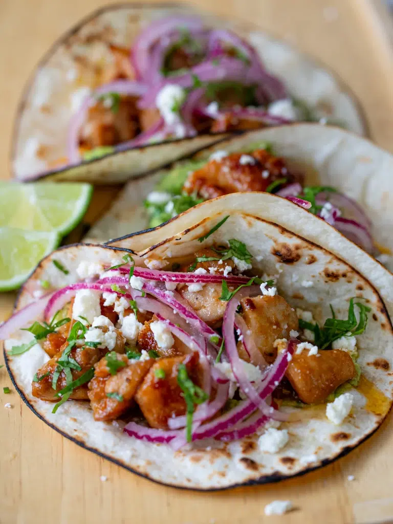 Sticky lime honey glazed chicken tacos topped with pickled red onions, crumbled cheese, and fresh herbs in toasted flour tortillas, served with lime wedges.